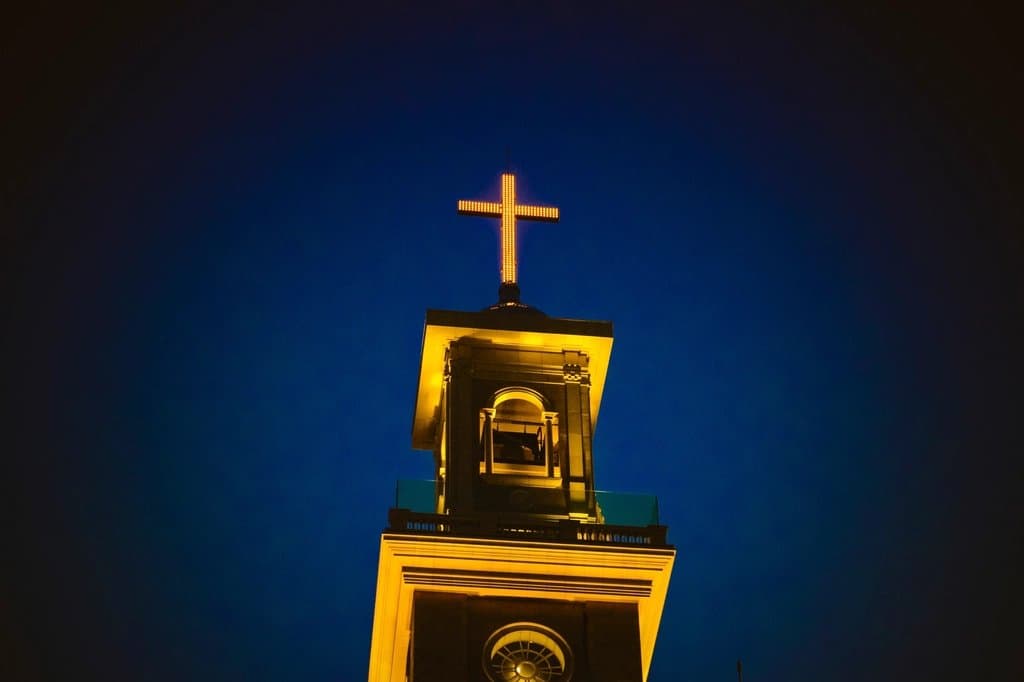 A lit church bell tower and cross against a deep blue evening sky